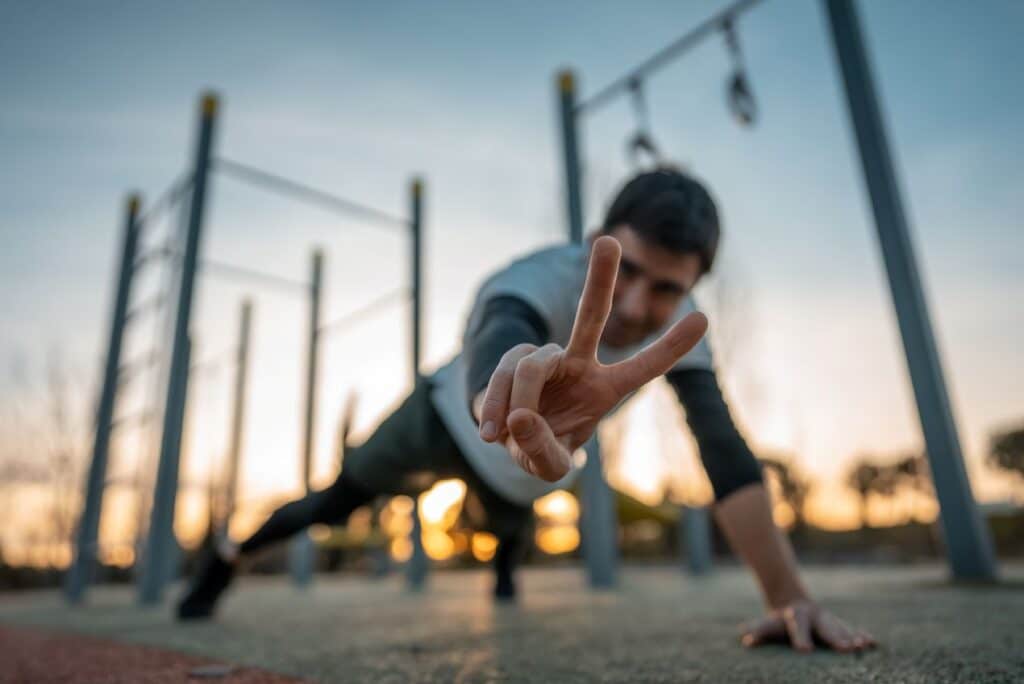 Sportler zeigt Victory-Zeichen waehrend Calisthenics-Workout im Park bei Sonnenuntergang